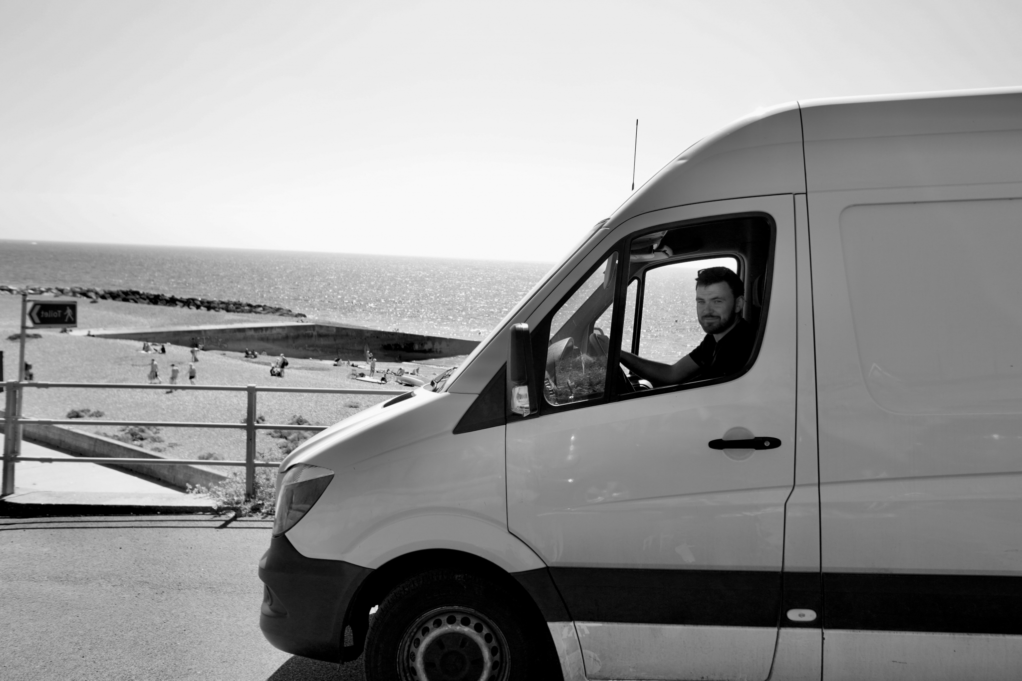 Peter Hawes at the wheel of the ESV Removals Sprinter parked above the beach and groynes on the coast east of Brighton, in black and white
