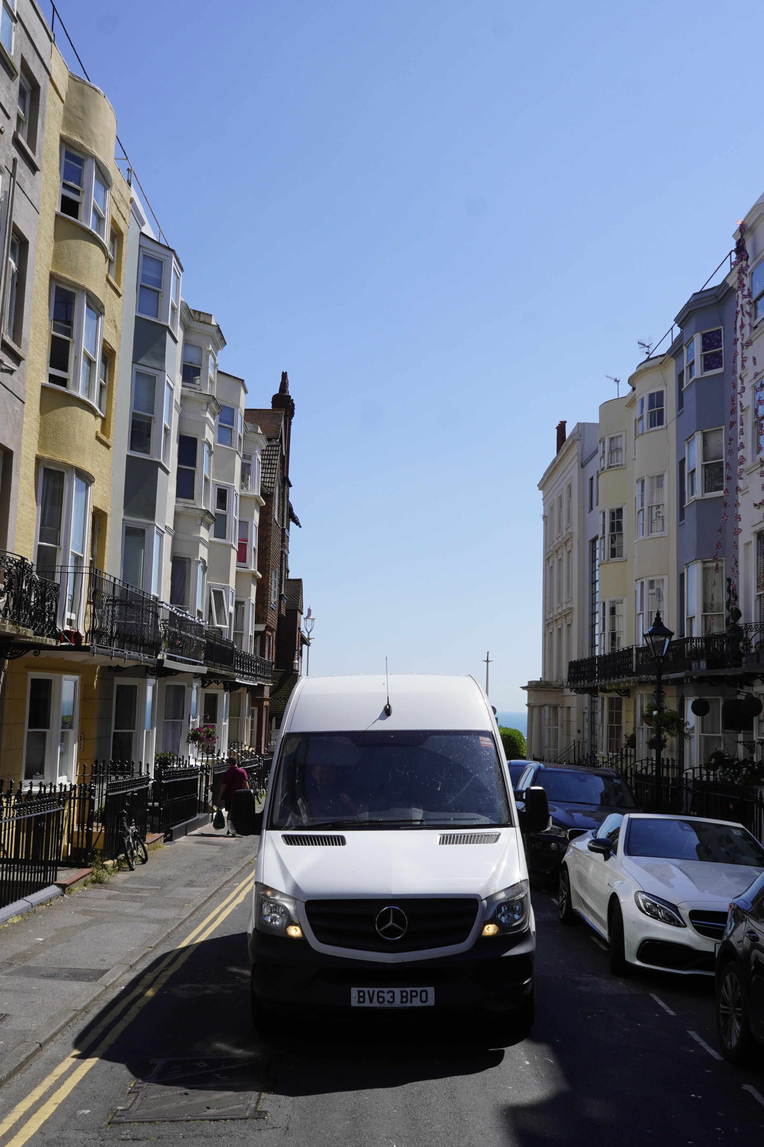 A white Mercedes Sprinter removals van parked head-on between cars on a narrow Kemptown street in Brighton, framed by balconied Regency townhouses with the sea visible at the end of the road.