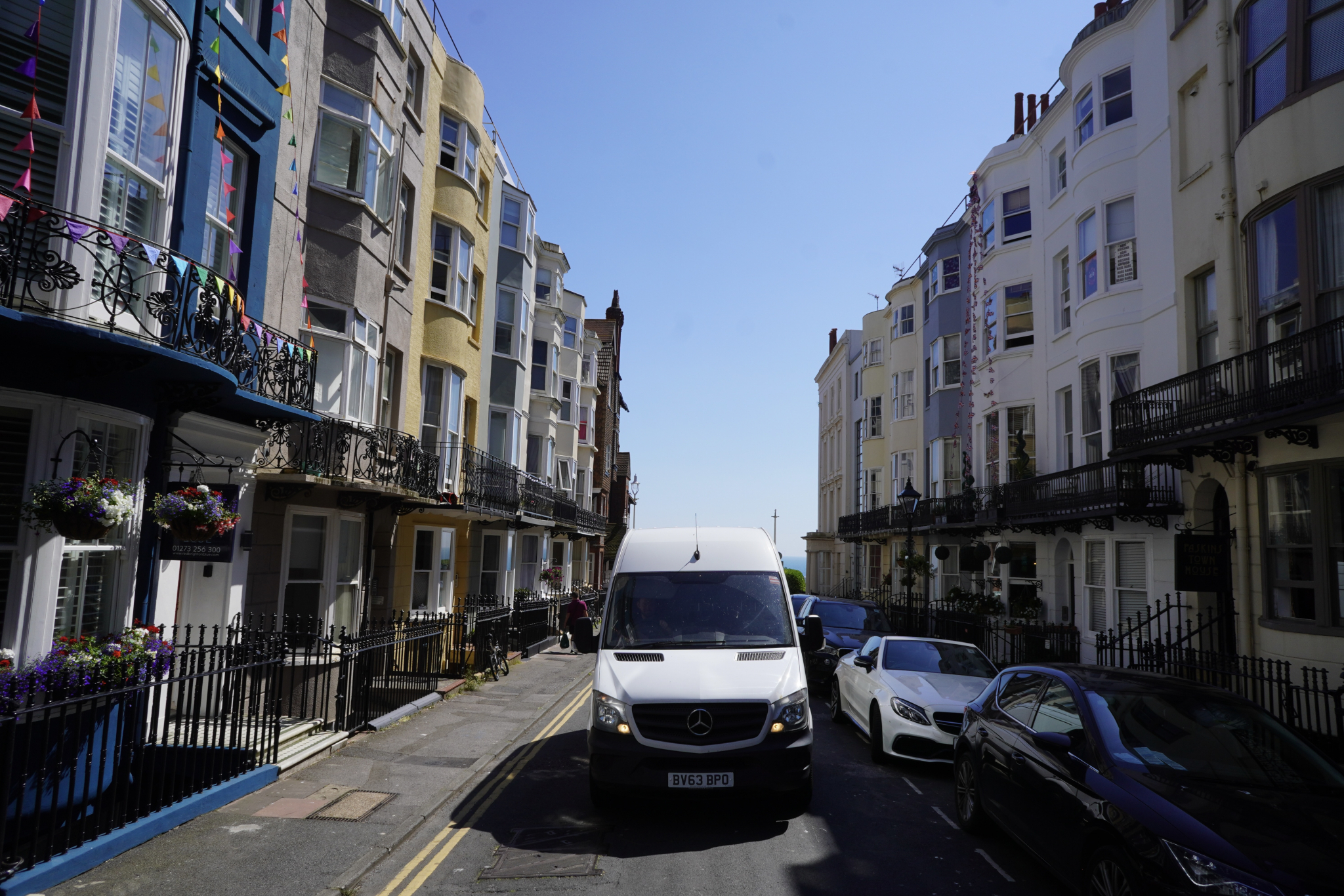 A white Mercedes Sprinter removals van driving down a narrow Kemptown street in Brighton, lined with Regency townhouses, wrought-iron balconies and double yellow lines, with the sea visible at the far end.