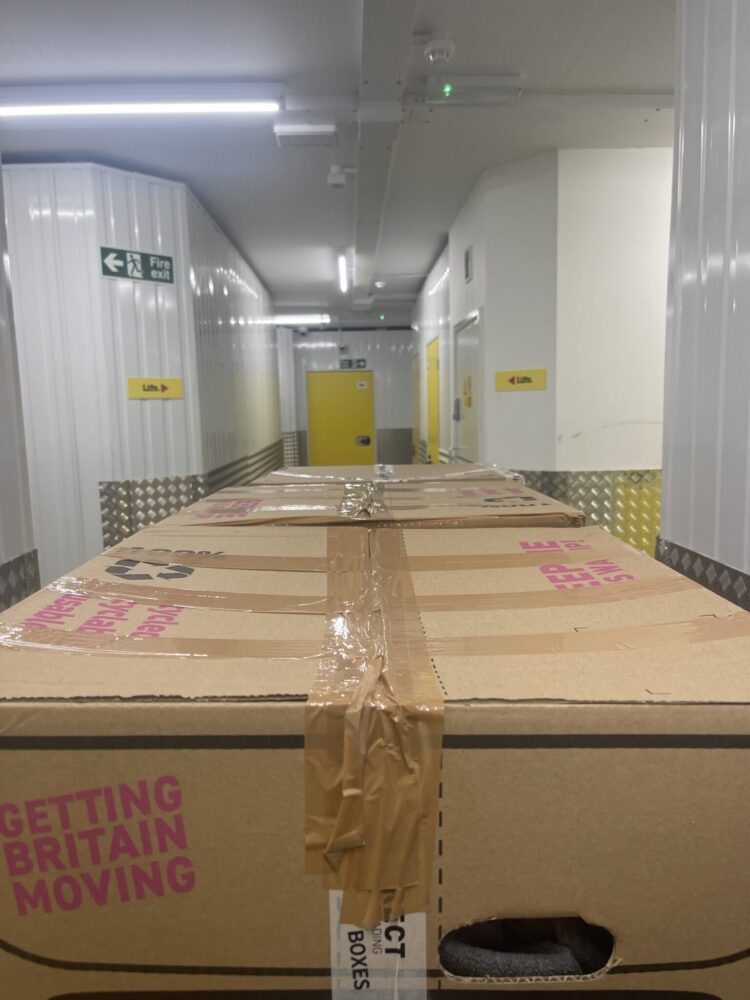 Taped cardboard moving boxes on a trolley in a storage corridor with lift signs and a fire exit, ready for a Brighton student move.