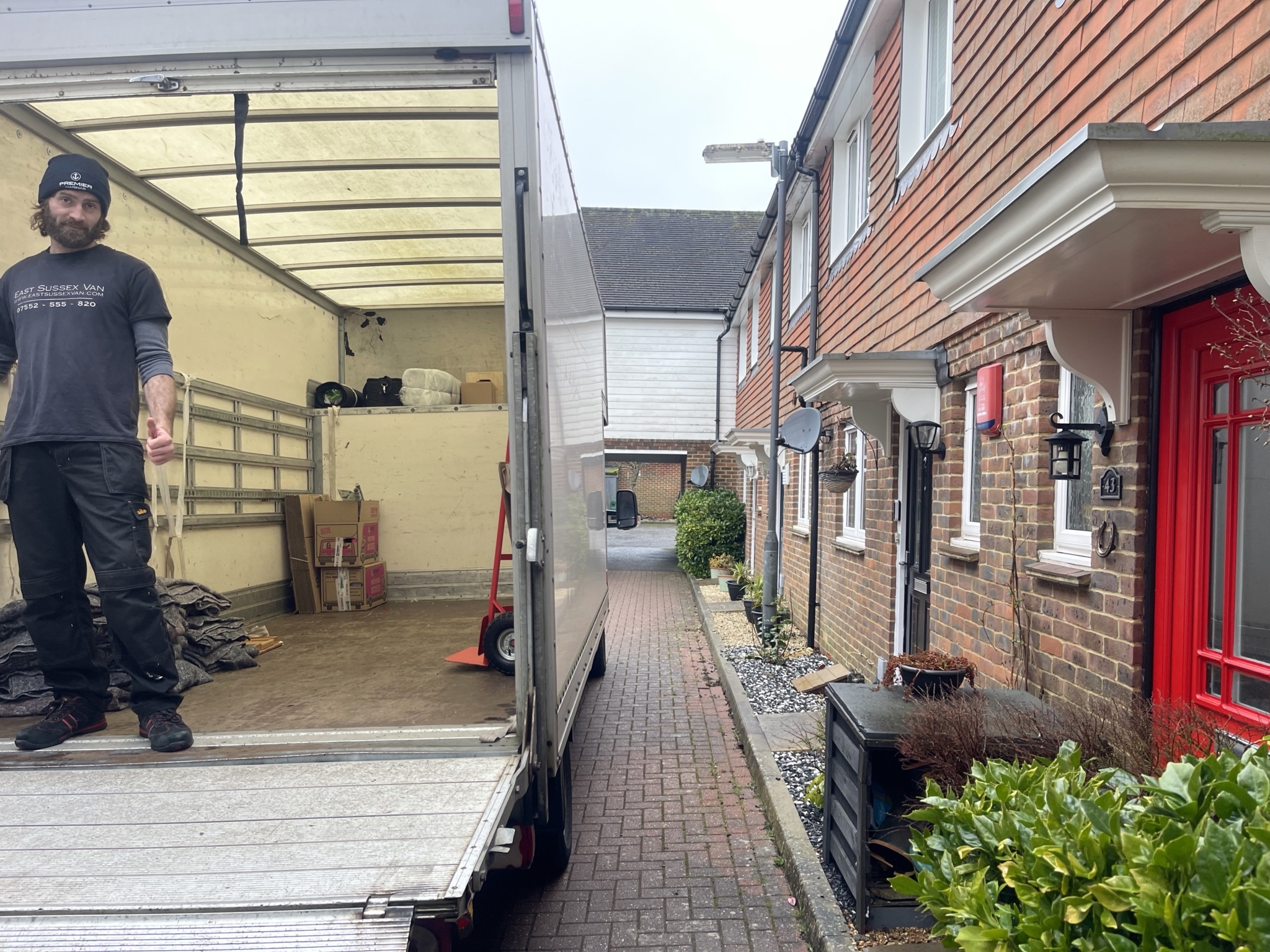 ESV Removals team member standing on the lowered ramp of a Luton van in a narrow residential lane beside brick and cladded houses, with moving blankets and boxes inside the van during a tight access move.
