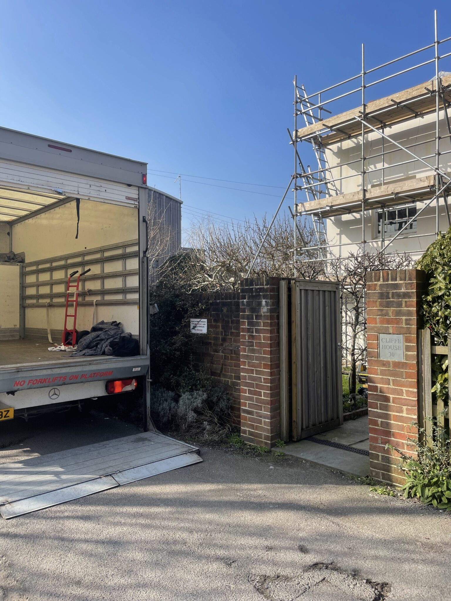 ESV Removals Luton van with tail lift lowered outside a brick gated entrance at a Lewes property, with scaffolding on the building and a hand truck inside the van ready for loading.