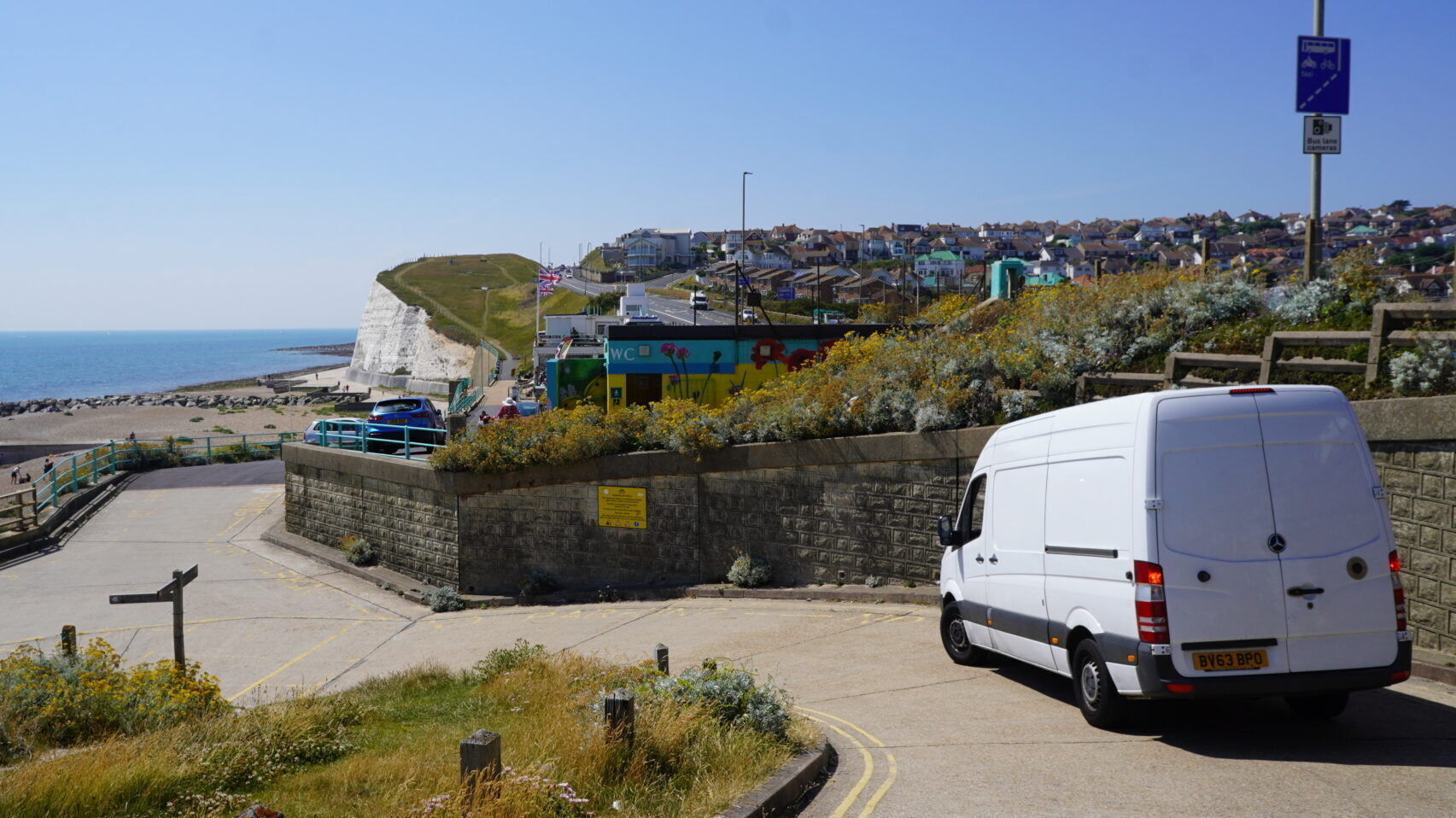 White removals van driving down a coastal road in Seaford, with the beach, sea, and white cliffs in the background on a clear day.
