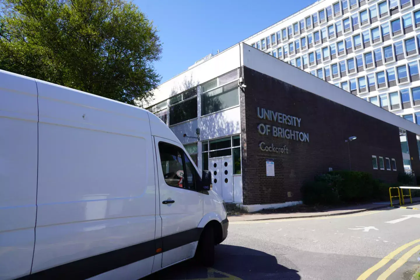 White removals van parked outside the University Of Brighton Cockcroft building on a clear day, with the campus signage visible on the wall.