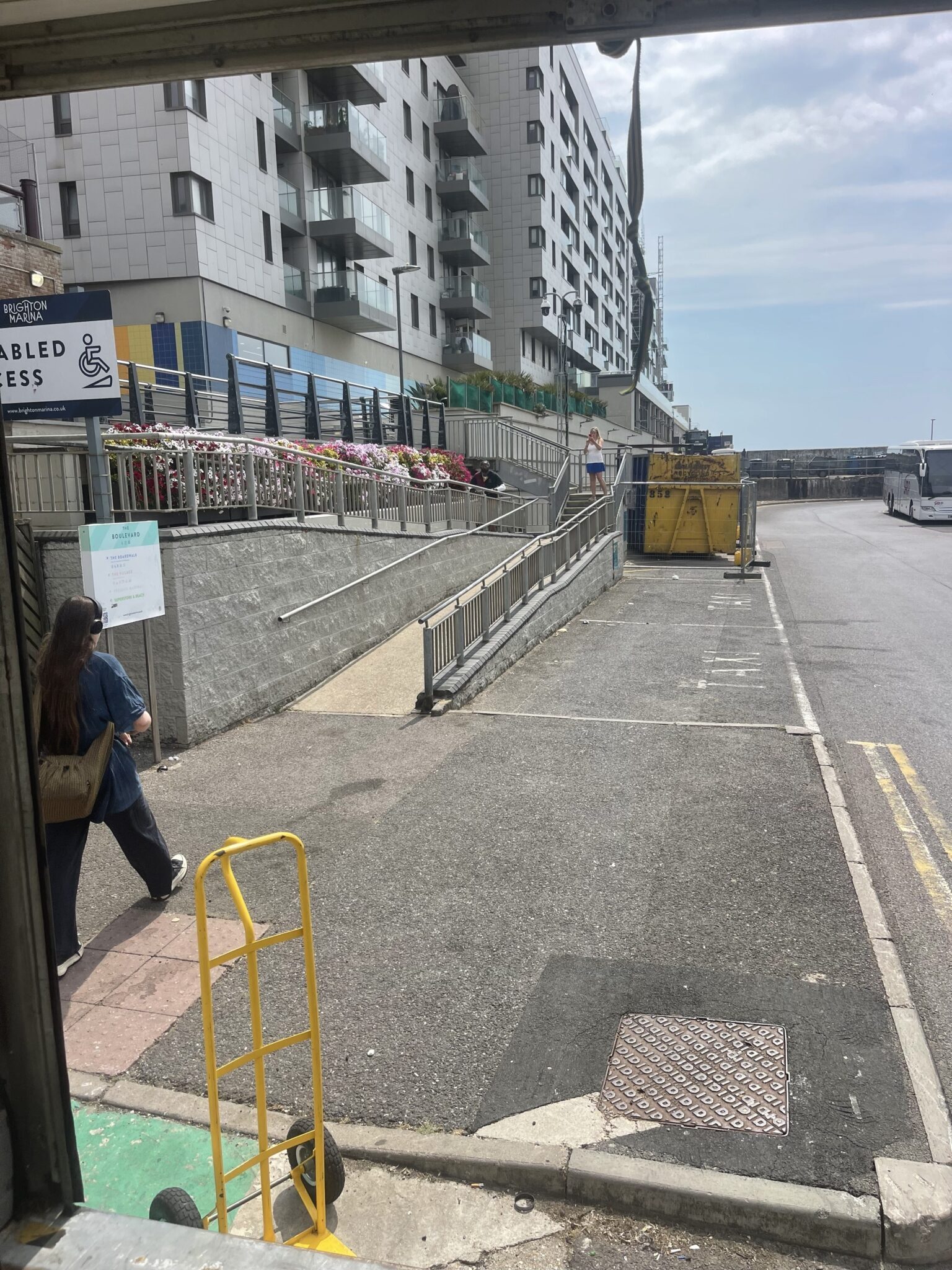 View from the back of an ESV Removals Luton van at Brighton Marina, a yellow sack truck ready on the loading area with a waterfront apartment tower, a skip behind Heras fencing and the Disabled Access ramp up to the boardwalk behind.
