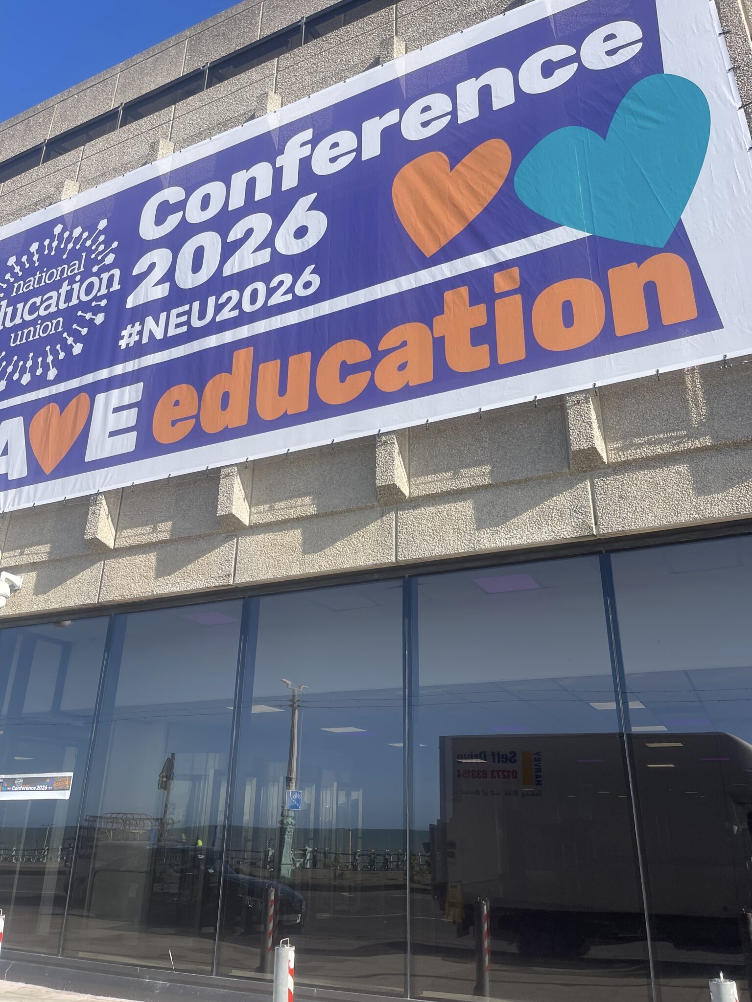 Large National Education Union Conference 2026 banner displayed outside Brighton Centre on the seafront, with the glass frontage reflecting the road, promenade and event vehicle.