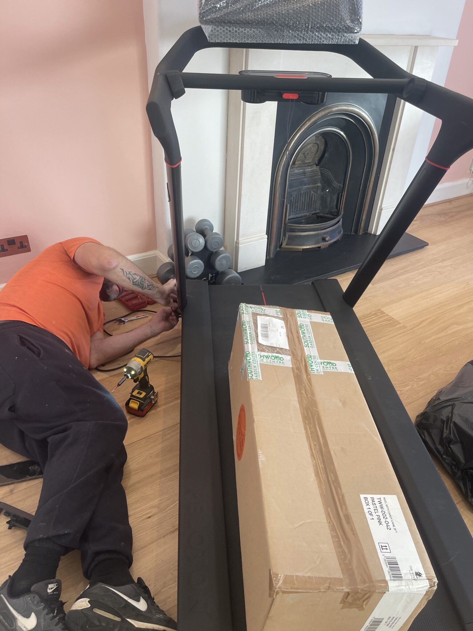 ESV Removals handyman from the snag list eliminator service lying on the floor with a drill, assembling a black treadmill in a Brighton living room with dumbbells and a fireplace in the background.