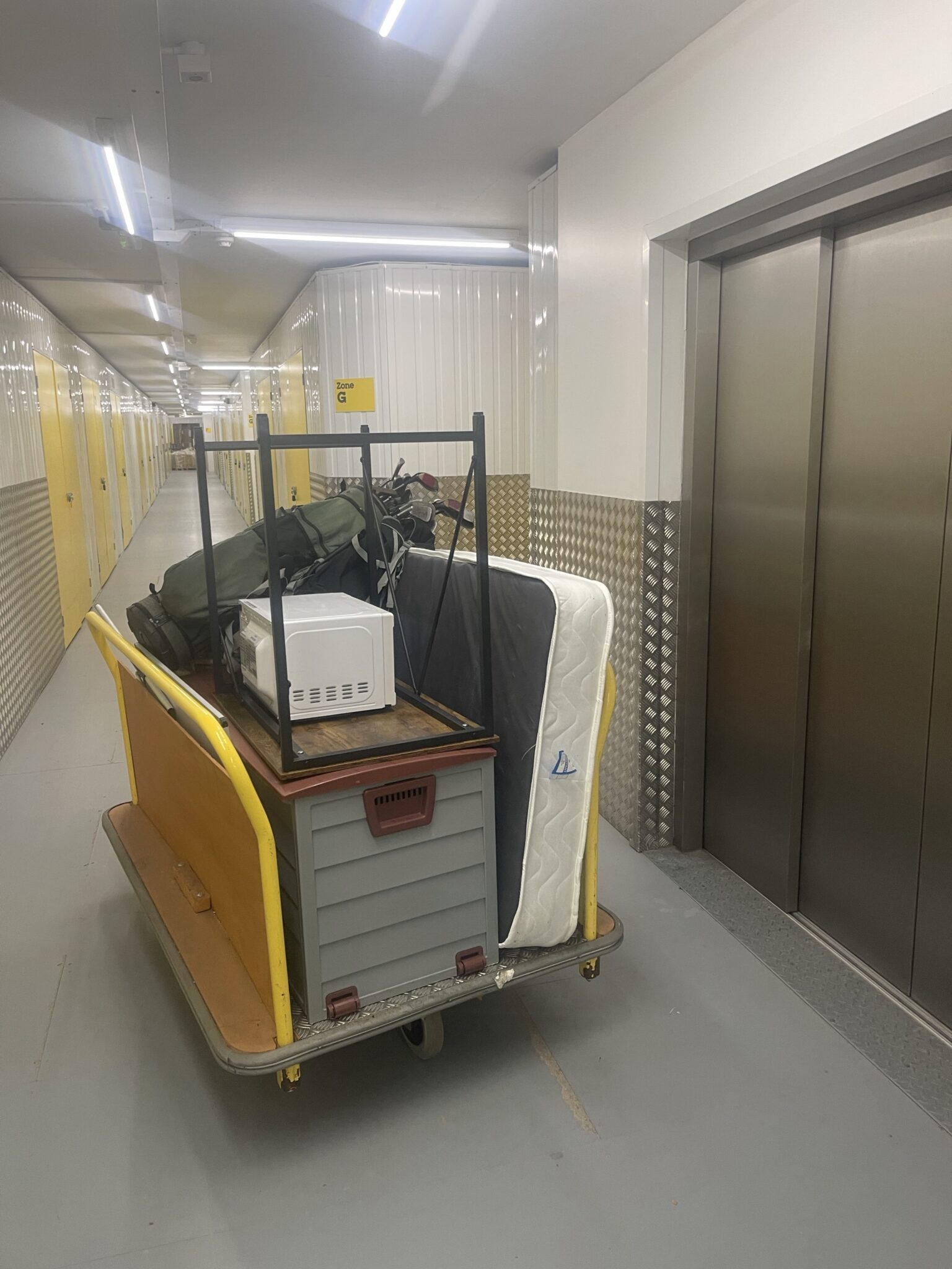 Yellow-handled trolley stacked with a mattress, golf clubs, microwave and plastic boxes beside a lift in a corridor at Big Yellow Self Storage Nottingham, during an ESV Removals Brighton man and van job moving a unit between sites.