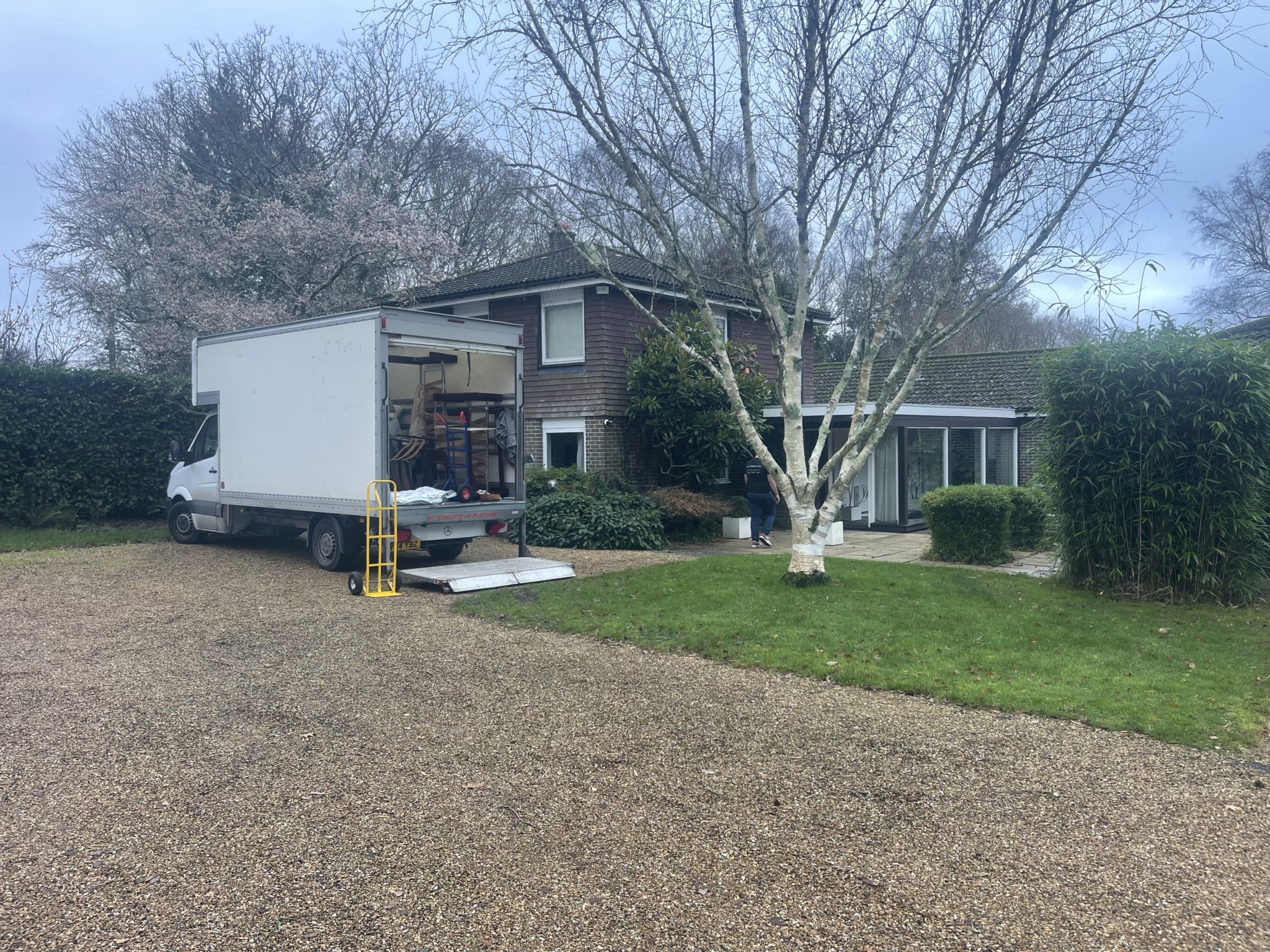 White Luton removals van with ramp and yellow sack barrow parked on a gravel driveway outside a detached house in Laughton, Sussex, with an ESV Removals team member walking to the front door during a house move.