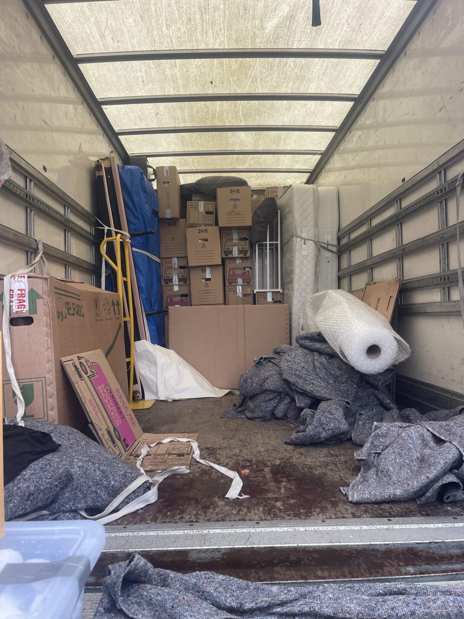 Inside a loaded ESV Removals Luton van, with boxes stacked tight at the front, tall flat items standing upright and strapped, and moving blankets and bubble wrap on the floor
