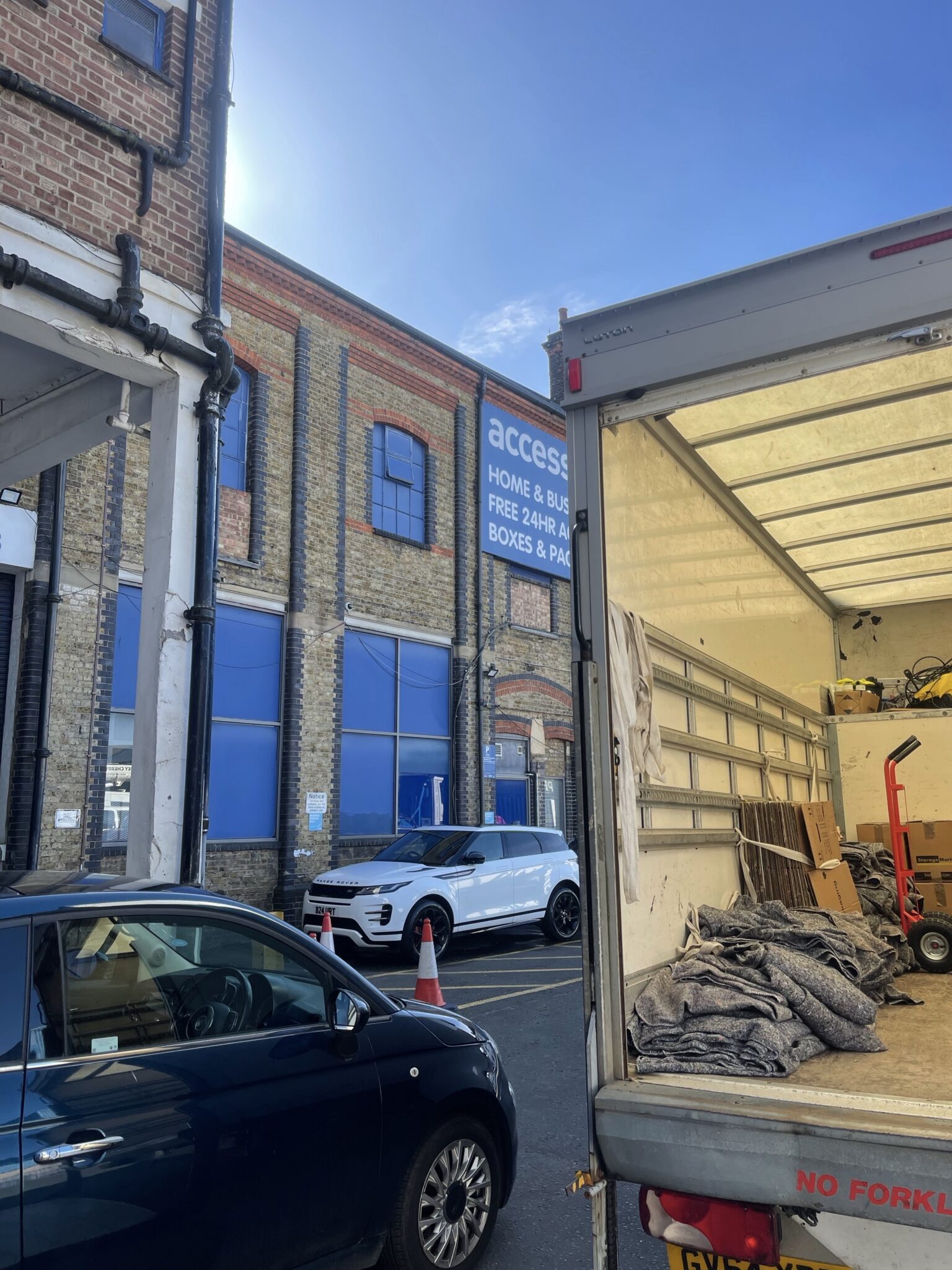Open rear of a Luton removals van being loaded outside a brick storage building, with moving blankets and moving equipment inside.