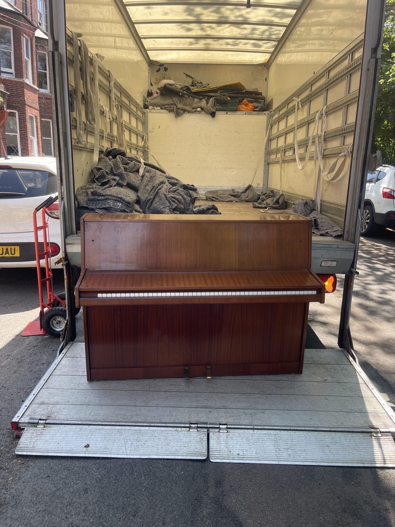 Upright piano on the tail lift of a Luton removals van, with moving blankets and equipment inside, during a Brighton and Sussex house move.
