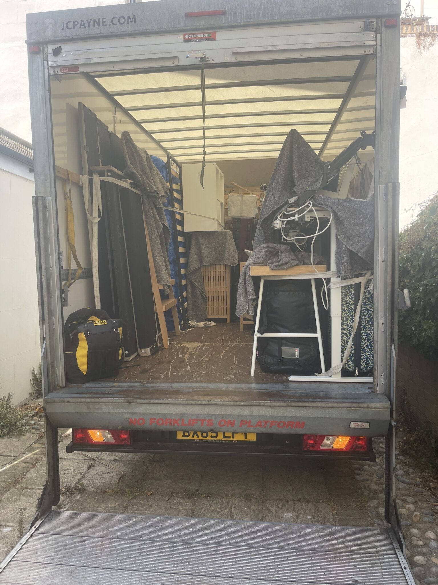 Rear view of a Luton removals van with the tail lift lowered, loaded with furniture wrapped in grey transport blankets, flat-packed panels and bed slats strapped to the side, a white desk frame with monitor arm and coiled cables, and a shelving unit secured with ratchet straps.