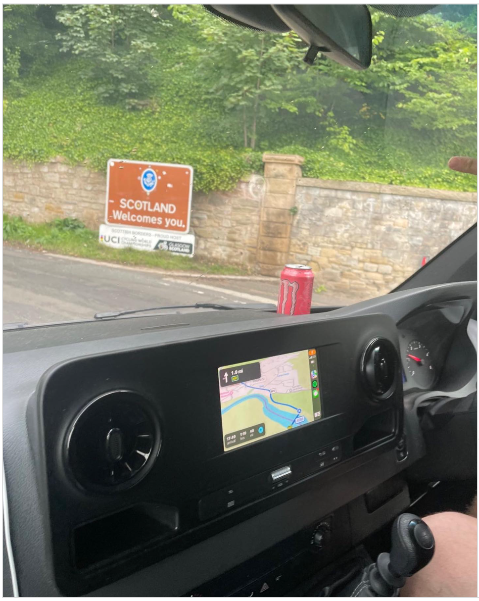 View from a van cab of the Scotland Welcomes You sign on a stone wall at the border, with the sat nav route showing on the dashboard