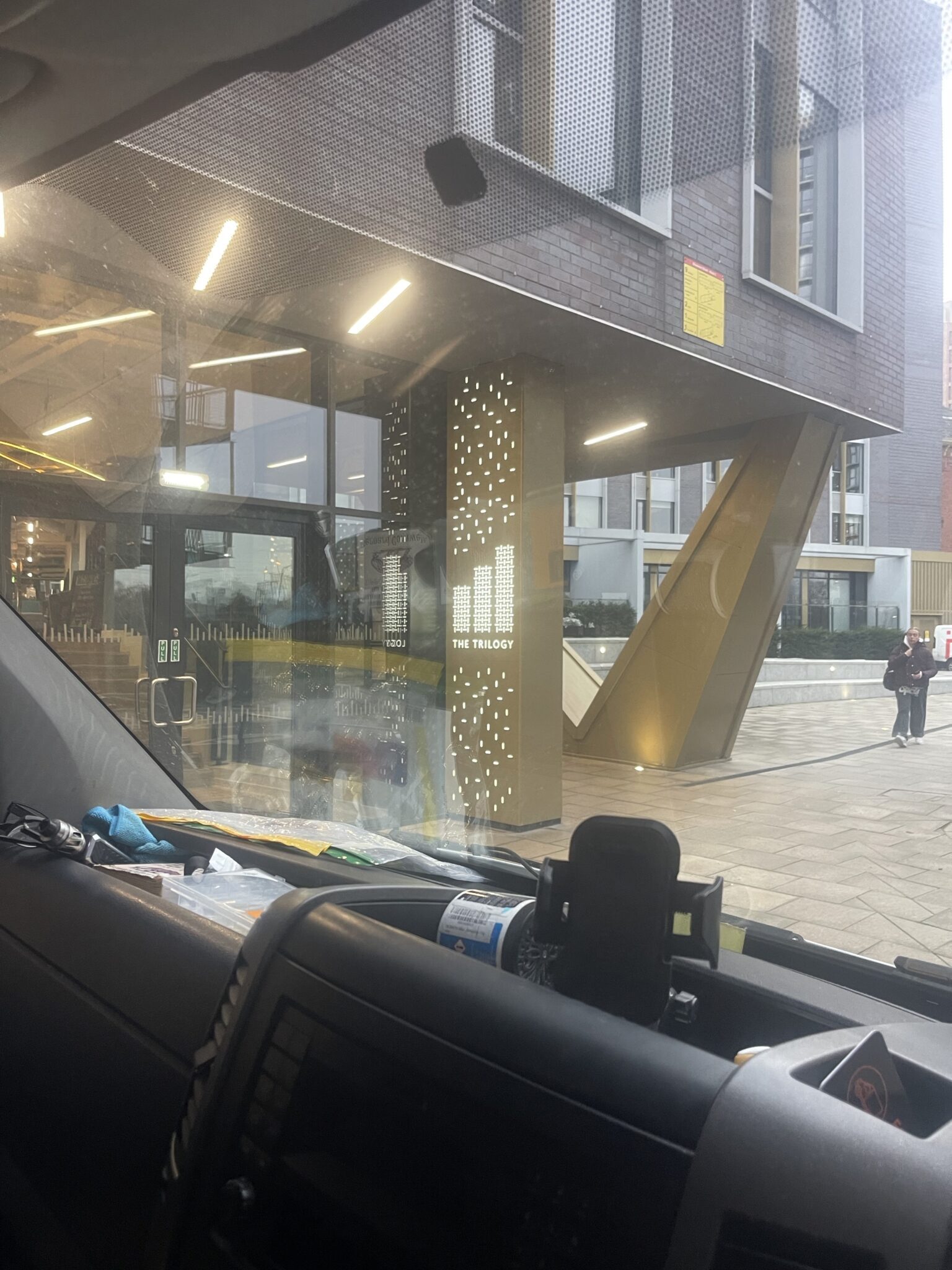 View from inside an ESV Removals van parked outside The Trilogy apartments in Manchester showing the illuminated entrance sign and modern building exterior before unloading a Brighton to Manchester house move.