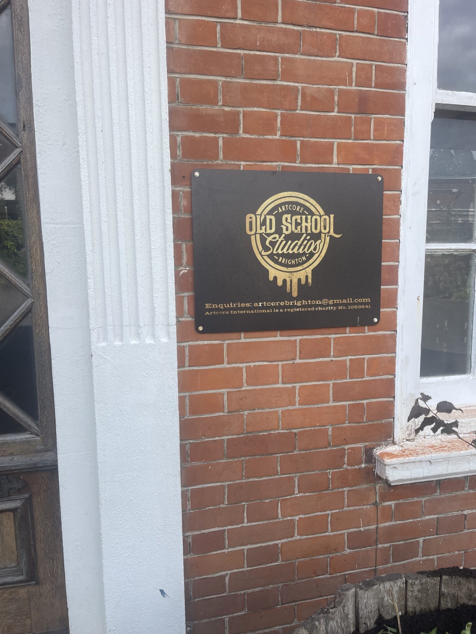 A black and gold metal sign reading 'Old School Studios, Artcore Brighton' mounted on a red brick wall beside a white-framed sash window, marking the entrance to the new art studio co-op the man and van move delivered to.