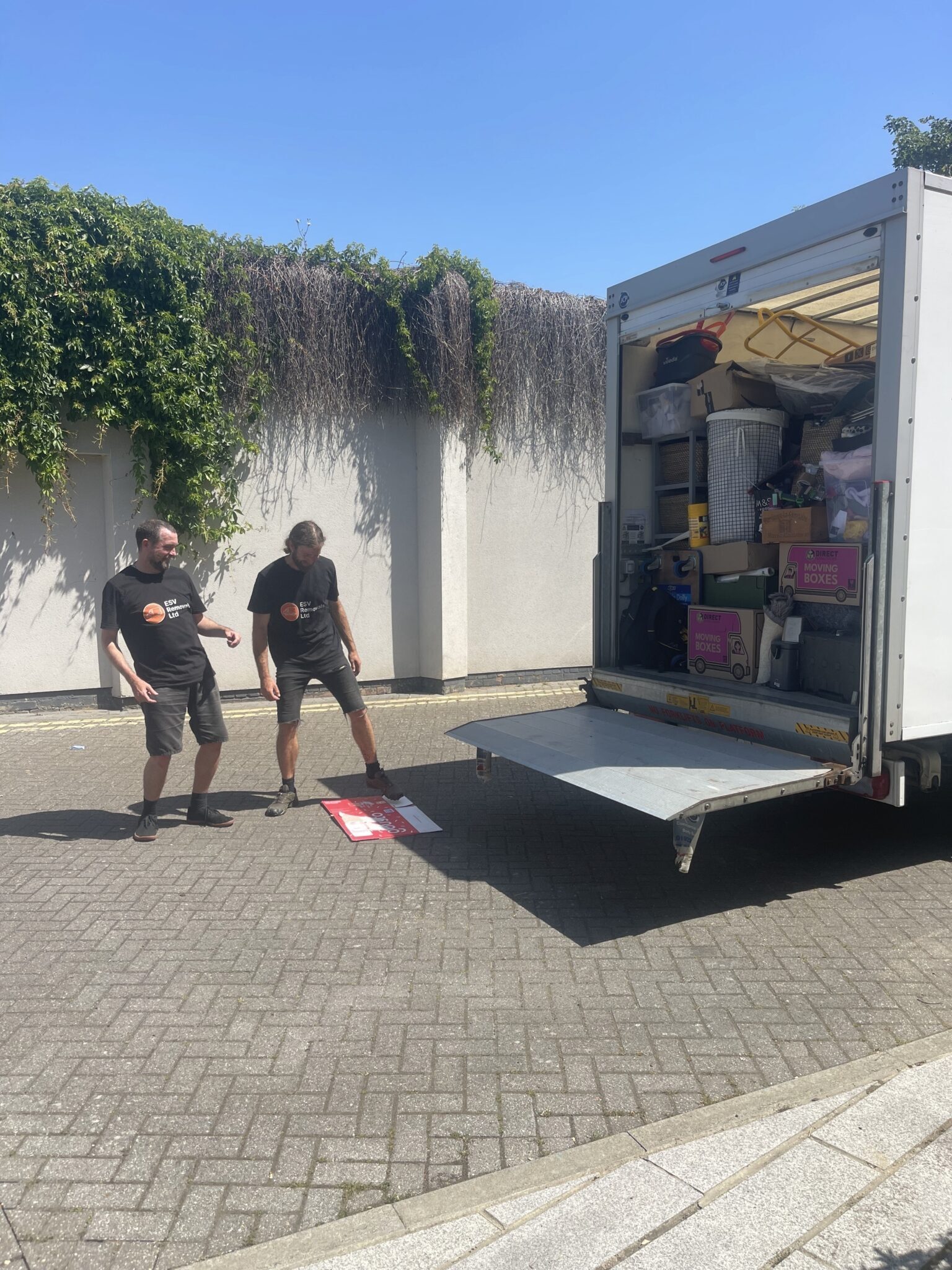 ESV Removals Ltd team in branded black t-shirts beside a fully loaded white Luton van with the tail lift lowered on a sunny day, the rear packed floor to ceiling with labelled moving boxes, plastic storage tubs, a laundry basket and household furniture during a full pack and move.