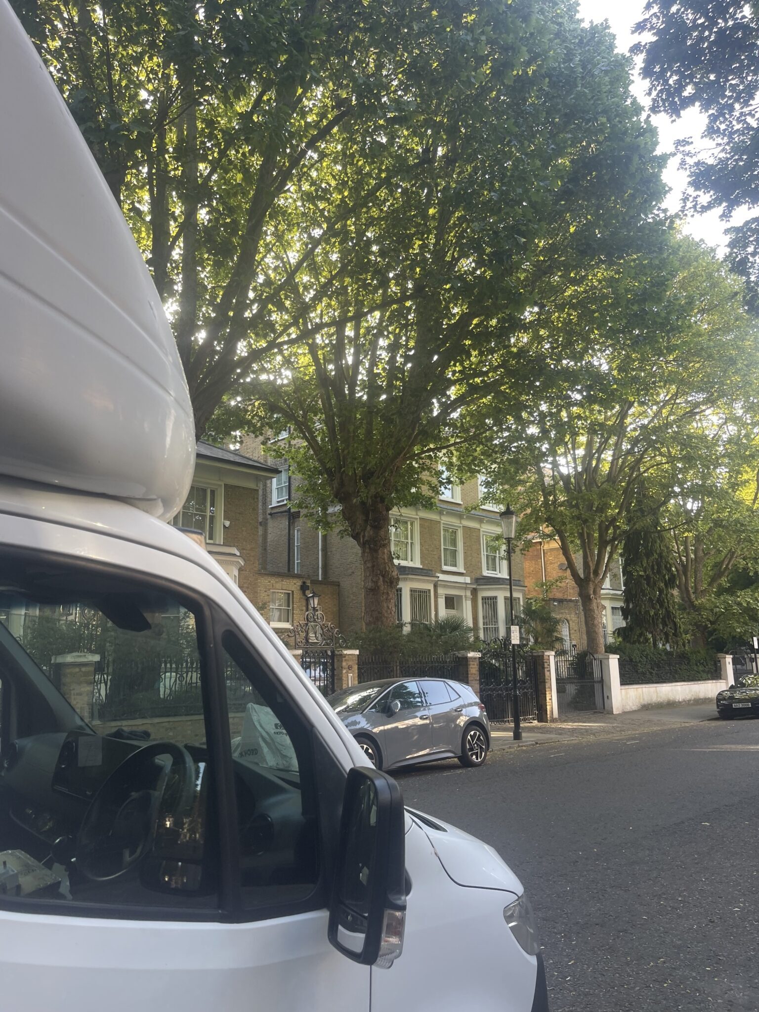 The front of ESV Removals' white Luton van parked on a leafy, tree-lined West London residential street of period brick terraces and iron railings, on the first delivery leg of the Kemptown to West London and Devon move.