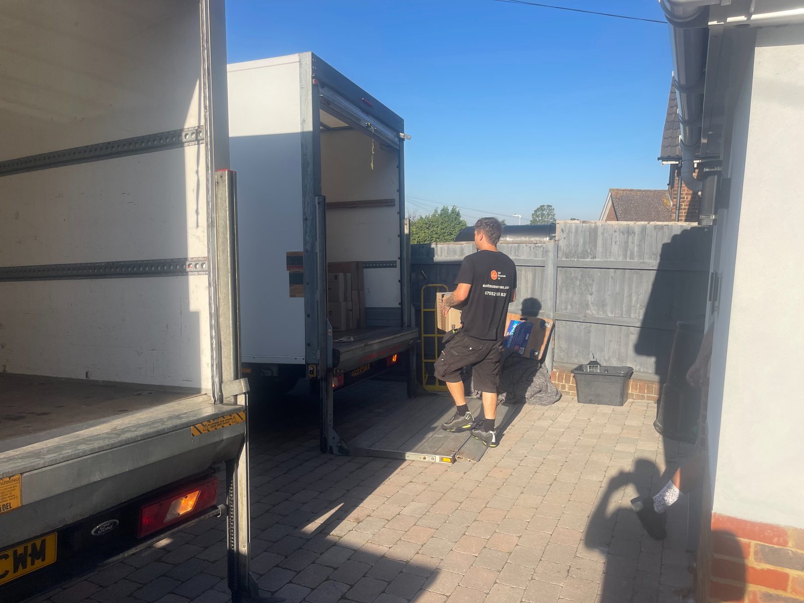 ESV Removals mover in a branded t-shirt carrying a moving box past a Luton van with its tail lift lowered