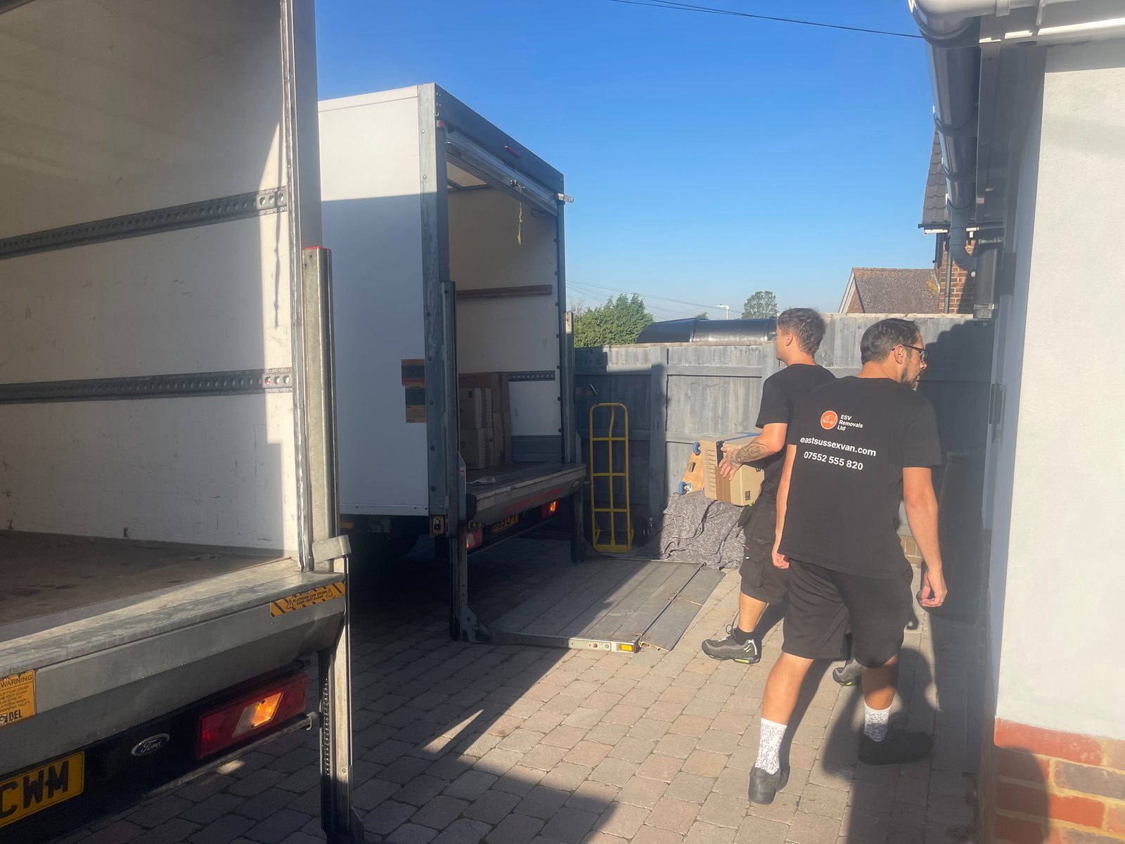 Two ESV Removals Luton vans with their tail lifts down on a block paved driveway, with two crew members in branded t-shirts carrying moving boxes