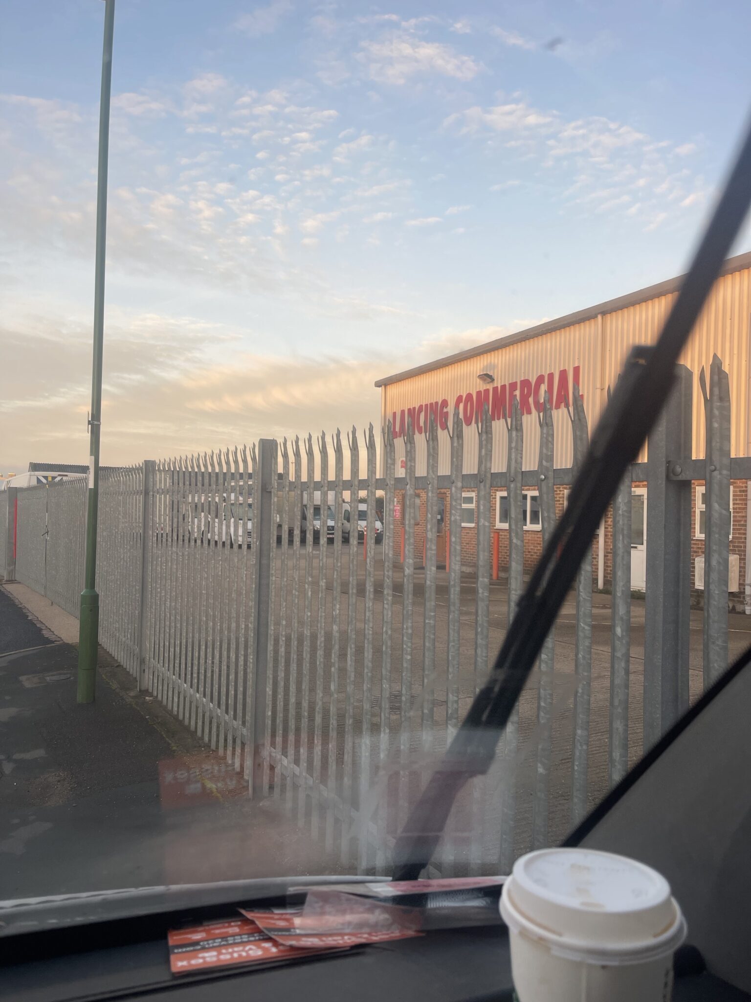 View through a van windscreen at dawn towards a commercial vehicle yard in Lancing behind a steel palisade fence, a coffee cup on the dashboard