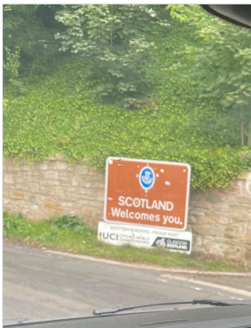 View from inside a vehicle approaching the Scotland border sign reading “Scotland Welcomes You,” beside a stone wall and greenery during a Brighton to Scotland move.