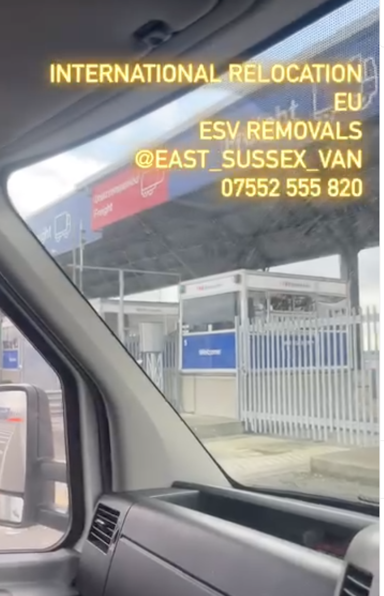 View from a removal van cab approaching a ferry terminal freight check-in booth, with signage and security gates ahead.