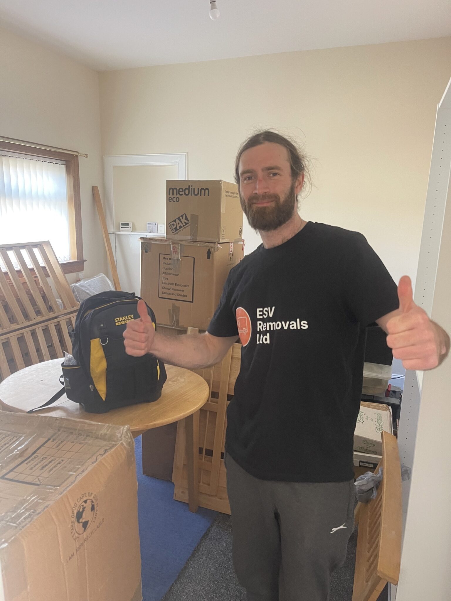 ESV Removals team member standing in a partly packed room with moving boxes, furniture and tools, giving a thumbs up during a home removals job.