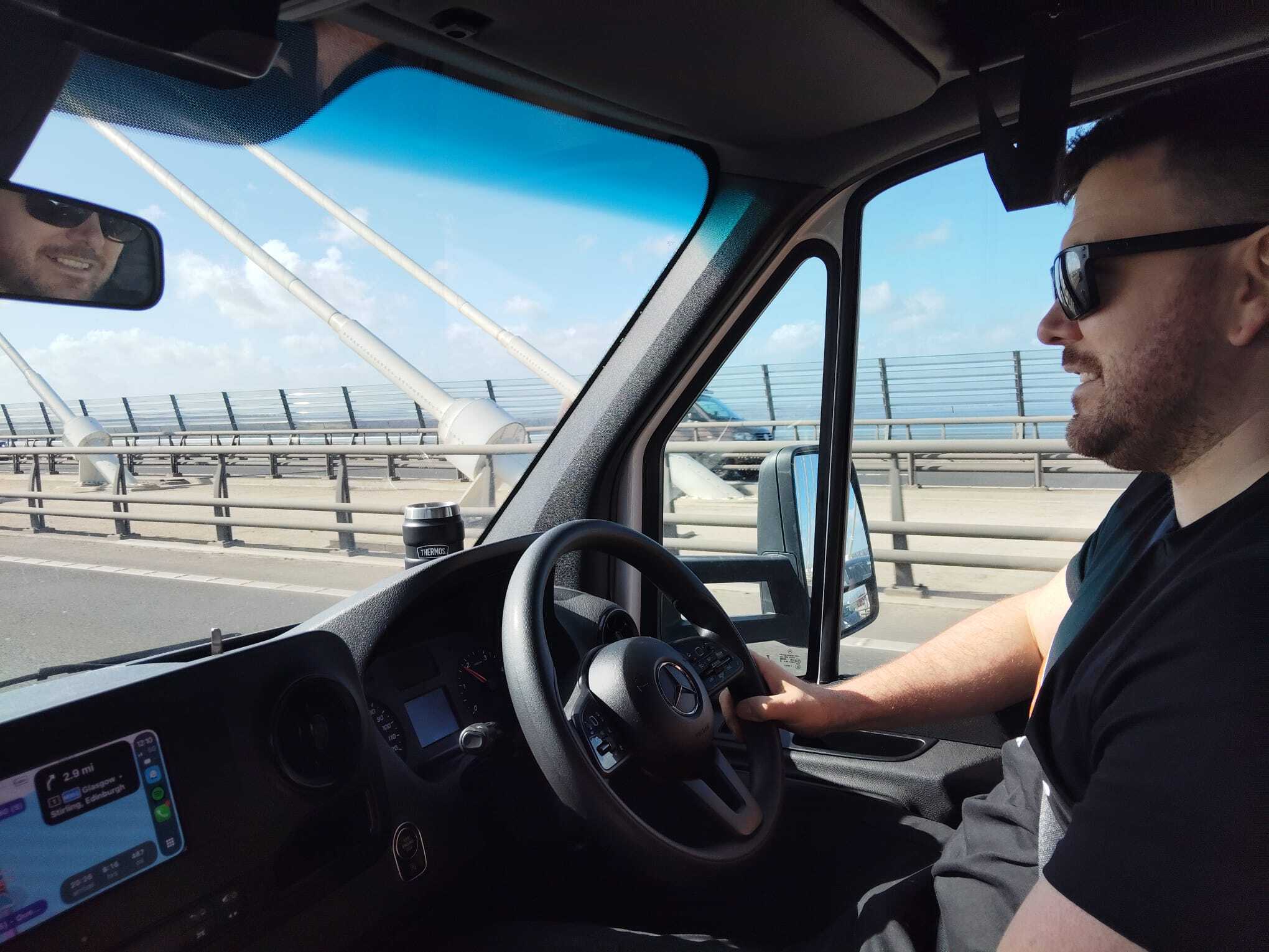 View from inside a Mercedes Sprinter as the driver crosses the Queen Elizabeth II Bridge on a long distance removals run, with blue skies, bridge cables and motorway barriers visible through the windscreen.