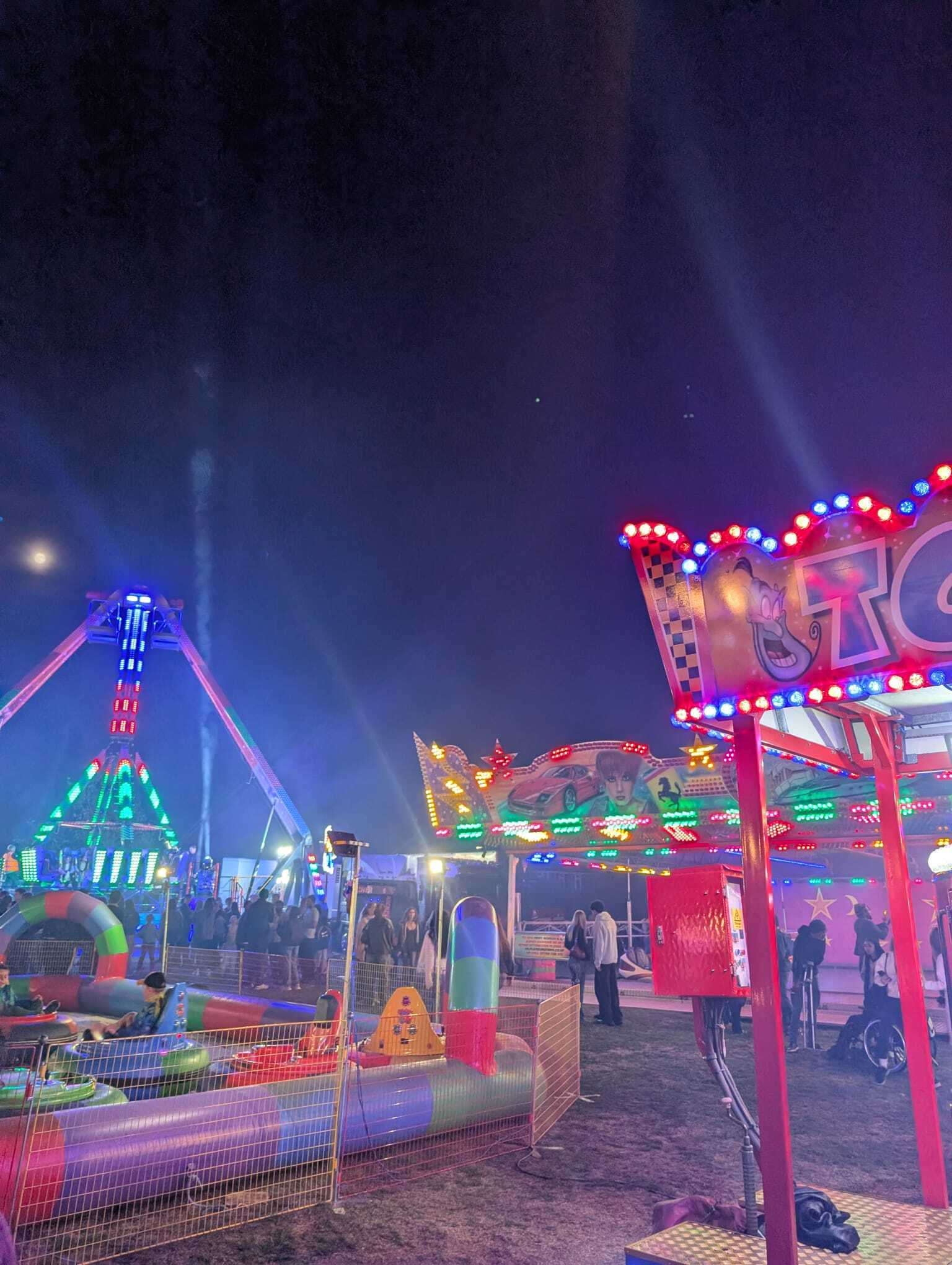 Brightly lit fairground rides at night, with crowds walking between them, at the Burgess Hill bonfire night event