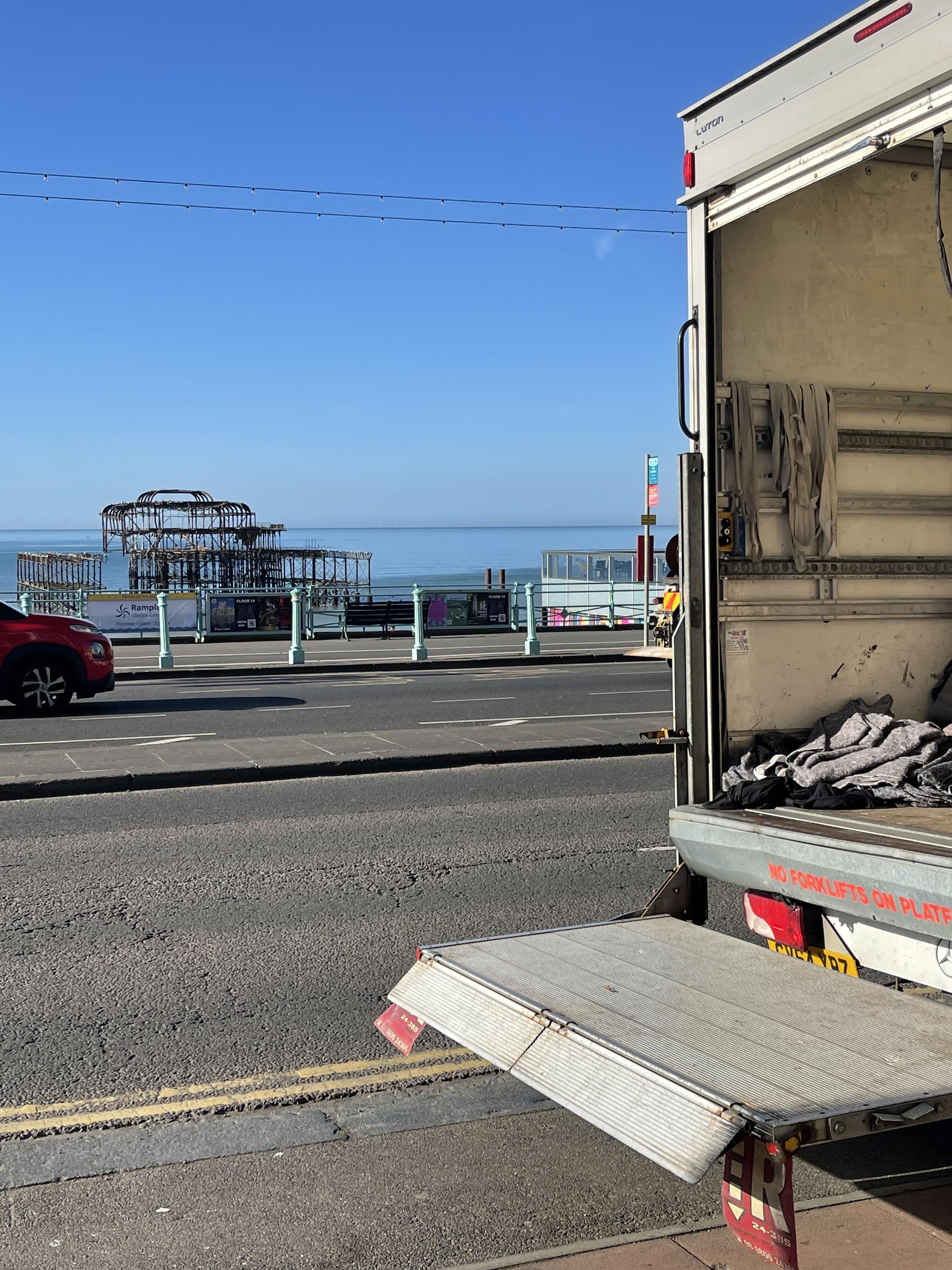 Open Luton removals van with tail lift down on Brighton seafront, with the West Pier remains visible across the road.