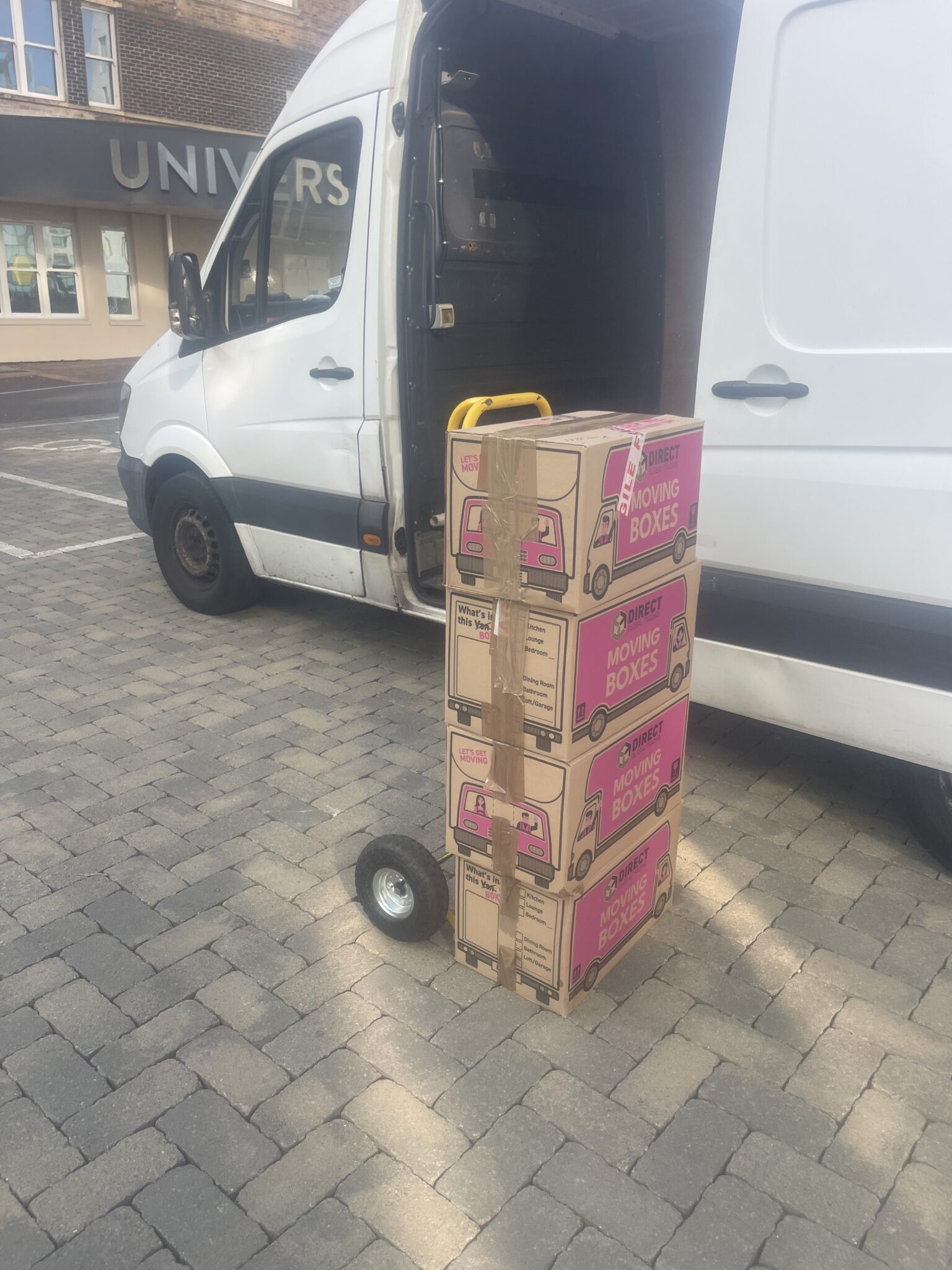 Four sealed moving boxes stacked on a sack truck beside the open side door of an ESV Removals Mercedes Sprinter van, with fragile tape visible on the top box during loading.