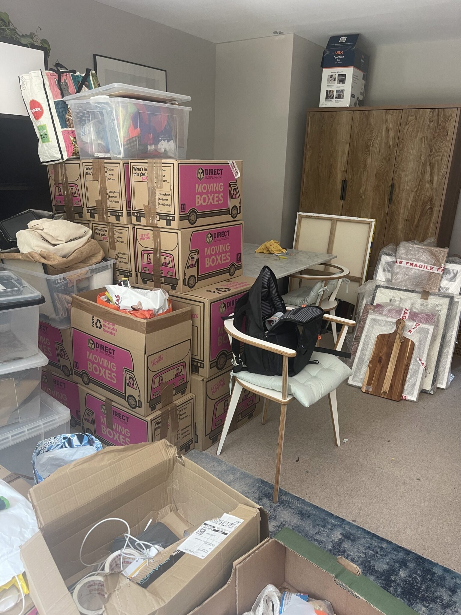 A living room in Peckham packed for moving day, with labelled cardboard boxes stacked by room and bubble-wrapped picture frames taped with fragile tape.