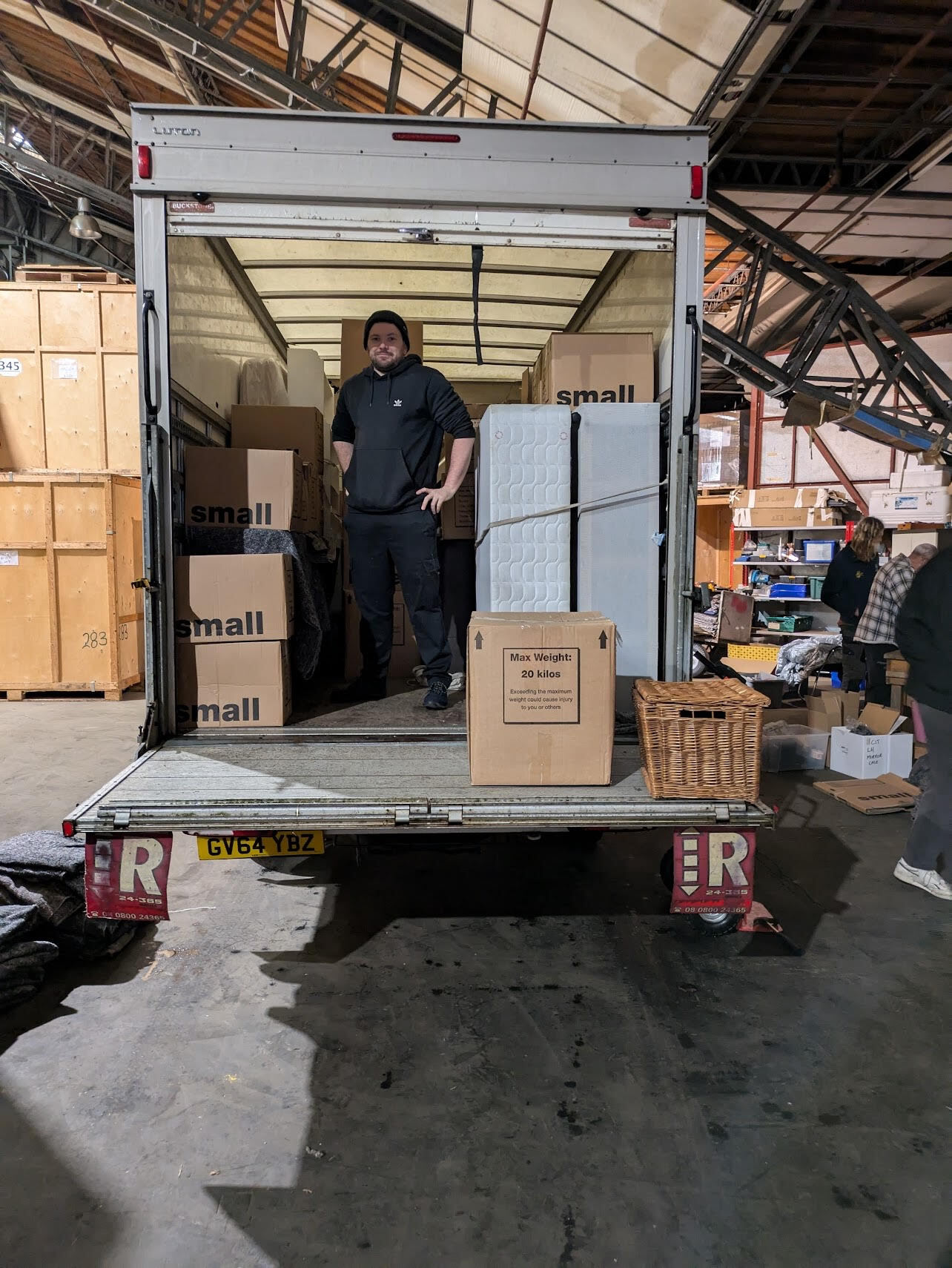 An ESV Removals crew member in a black hoodie and beanie standing in the back of a Luton van with the tail-lift down, part-loaded with boxes labelled 'small', a strapped-upright mattress and divan base, and a '20 kilos max' box, inside a storage warehouse with wooden storage crates behind.