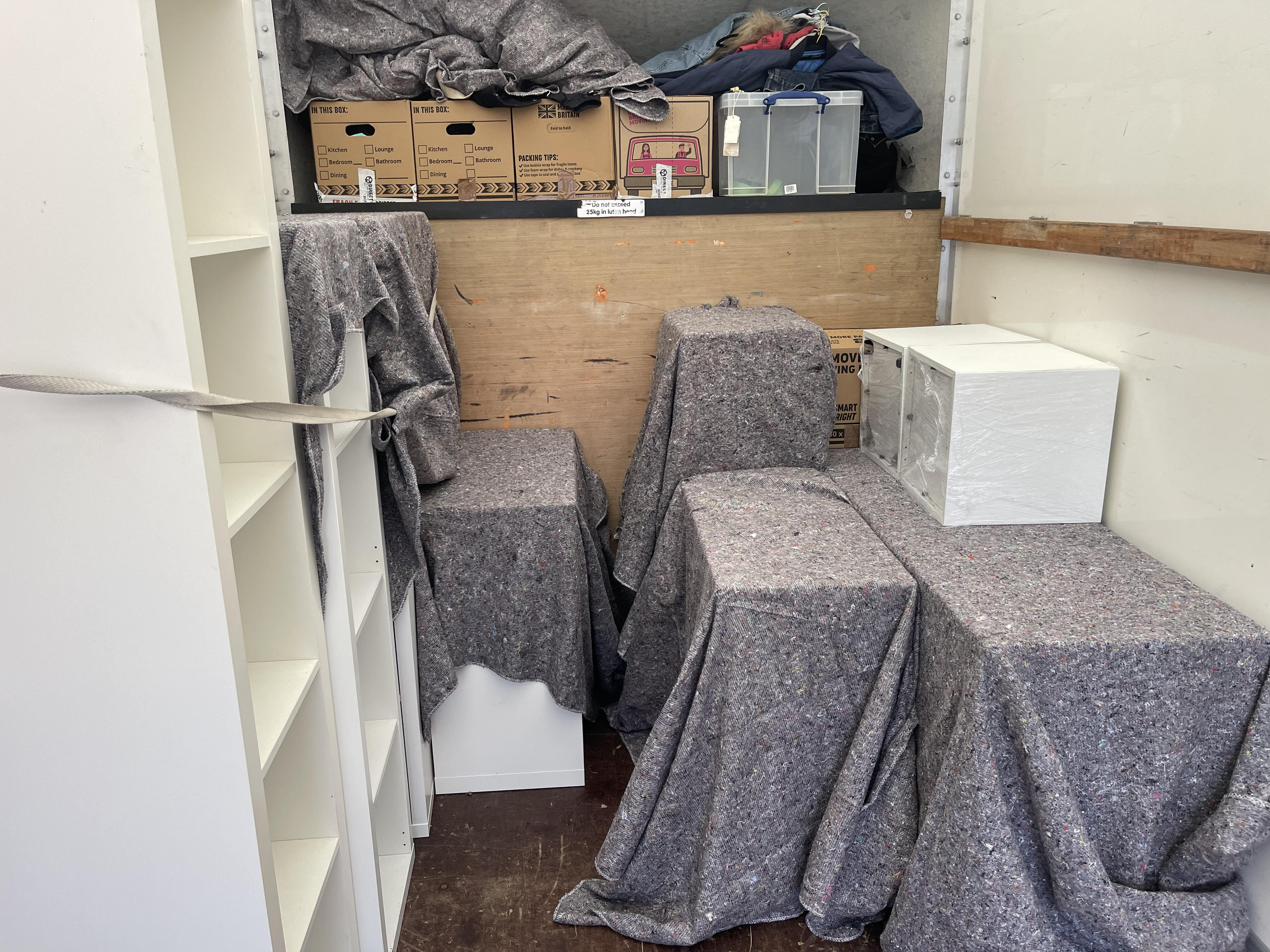Inside the removals van with furniture protected in grey moving blankets and secured with a strap, including a white shelving unit, covered items and a wrapped box, with labelled moving boxes and plastic storage tubs stacked at the front.