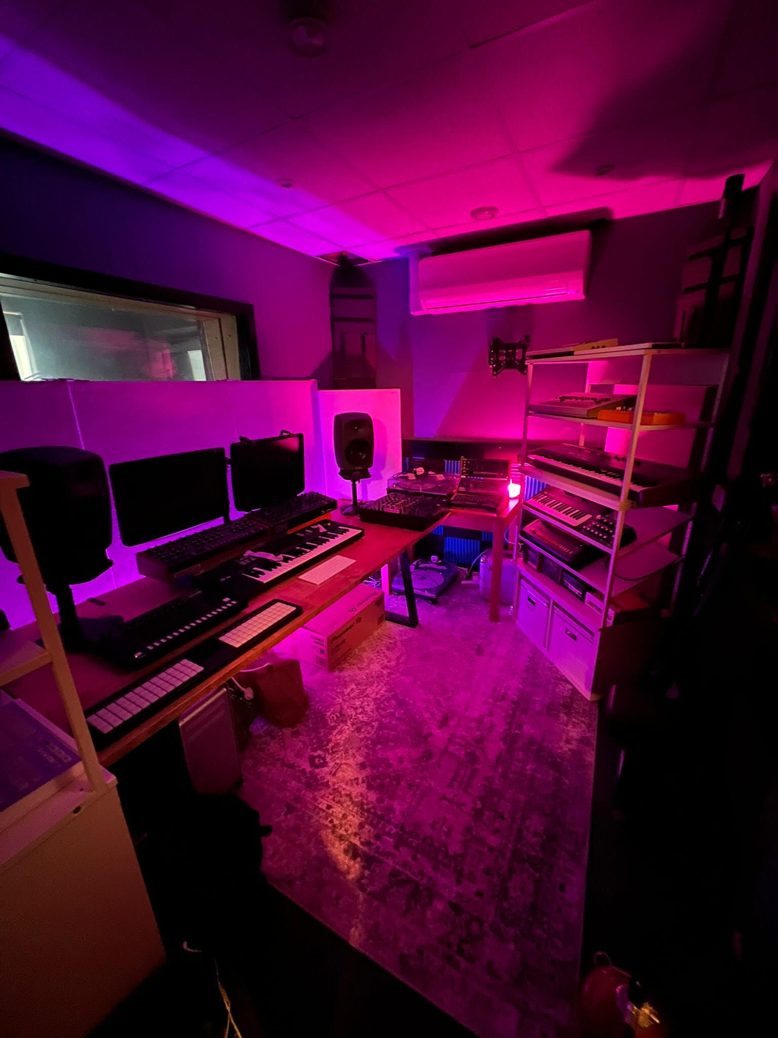 Music production studio reassembled after a move, with synthesisers, studio monitors, a mixing desk and keyboards arranged on desks and shelving under pink lighting.